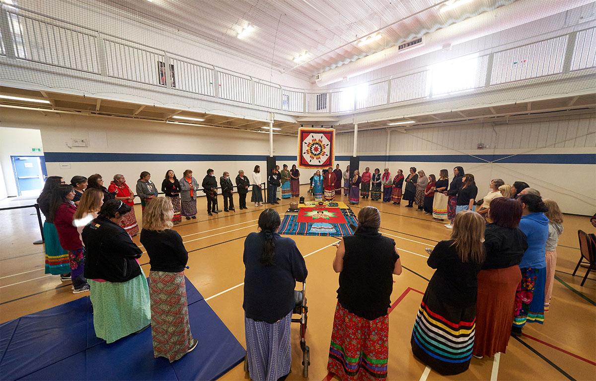 A wide-angle shot of a group of women, heads bowed and standing in a circle as part of an Indigenous water ceremony. In the middle of the circle, a woman stands in front of a blanket and mats.